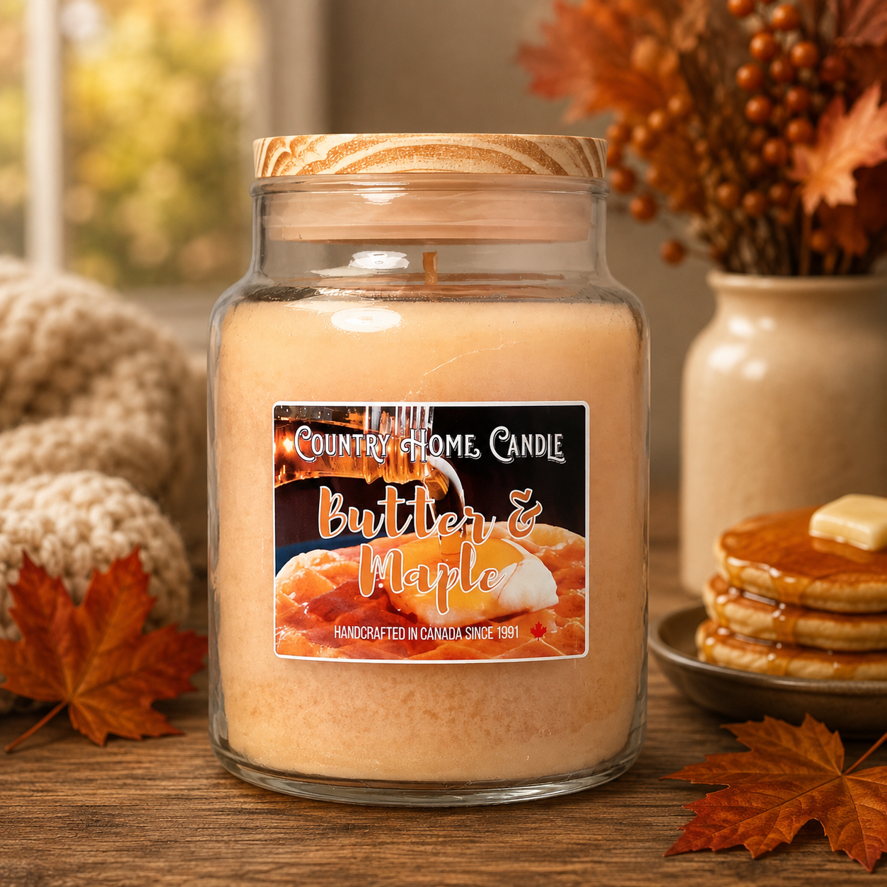 A large glass jar candle with a wooden lid sits on a wooden table, labeled 'Country Home Candle Butter & Maple.' The scene is styled with autumn leaves, a knitted blanket, pancakes, and a vase of fall flowers in the background.