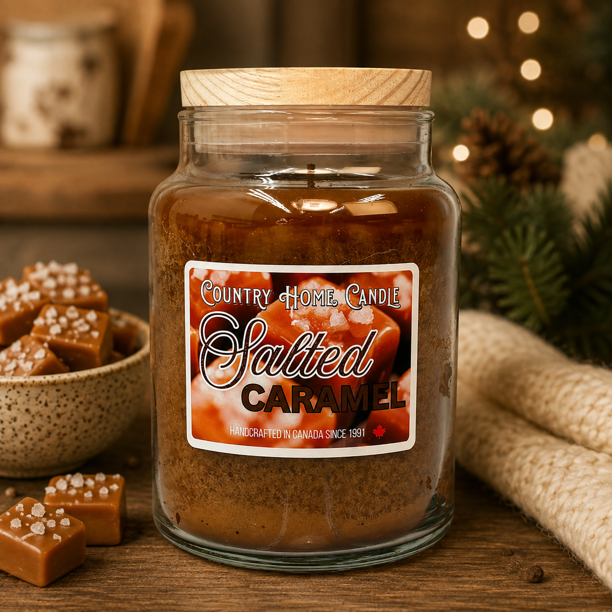 A large glass jar candle labeled 'Salted Caramel' by Country Home Candle, filled with brown wax and topped with a light wood lid, set on a wooden surface with caramel pieces and holiday decor in the background.
