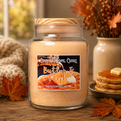 A large glass jar candle with a wooden lid sits on a wooden table, labeled 'Country Home Candle Butter & Maple.' The scene is styled with autumn leaves, a knitted blanket, pancakes, and a vase of fall flowers in the background.