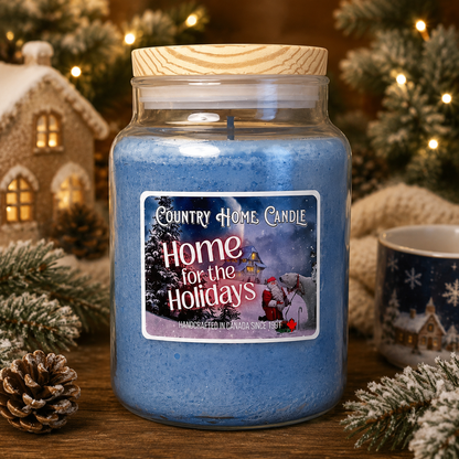 A blue scented candle in a clear glass jar with a light wood lid, displayed on a wooden surface with holiday decorations including a miniature house, pine branches, and a mug. The label reads 'Country Home Candle Home for the Holidays'.