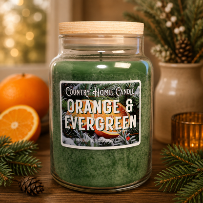 A large green scented candle in a clear glass jar with a light wood lid, labeled 'Country Home Candle Orange & Evergreen,' styled on a wooden surface with oranges, pine branches, and a warm blurred holiday background.