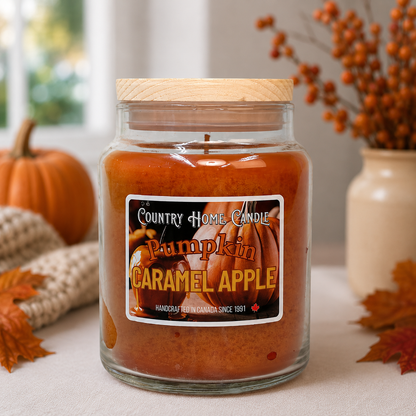 A large glass jar candle with a wooden lid and a front label reading 'Country Home Candle Pumpkin Caramel Apple,' styled on a table with pumpkins, autumn leaves, and a vase of dried berries in the background.